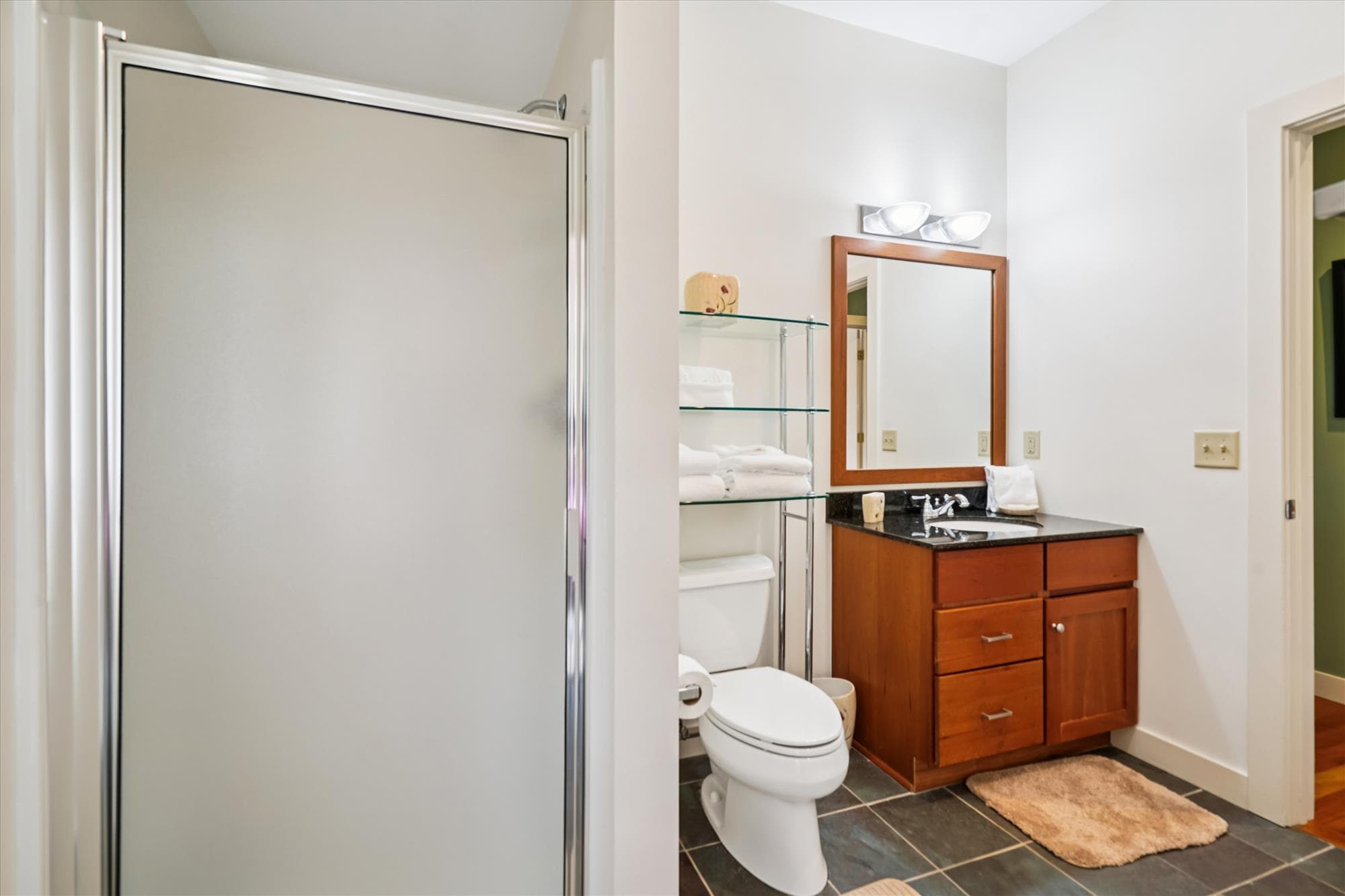 A bright full bath featuring a walk-in shower, granite vanity, and glass shelving stocked with fresh towels. Conveniently located near the queen bedroom for privacy and comfort.