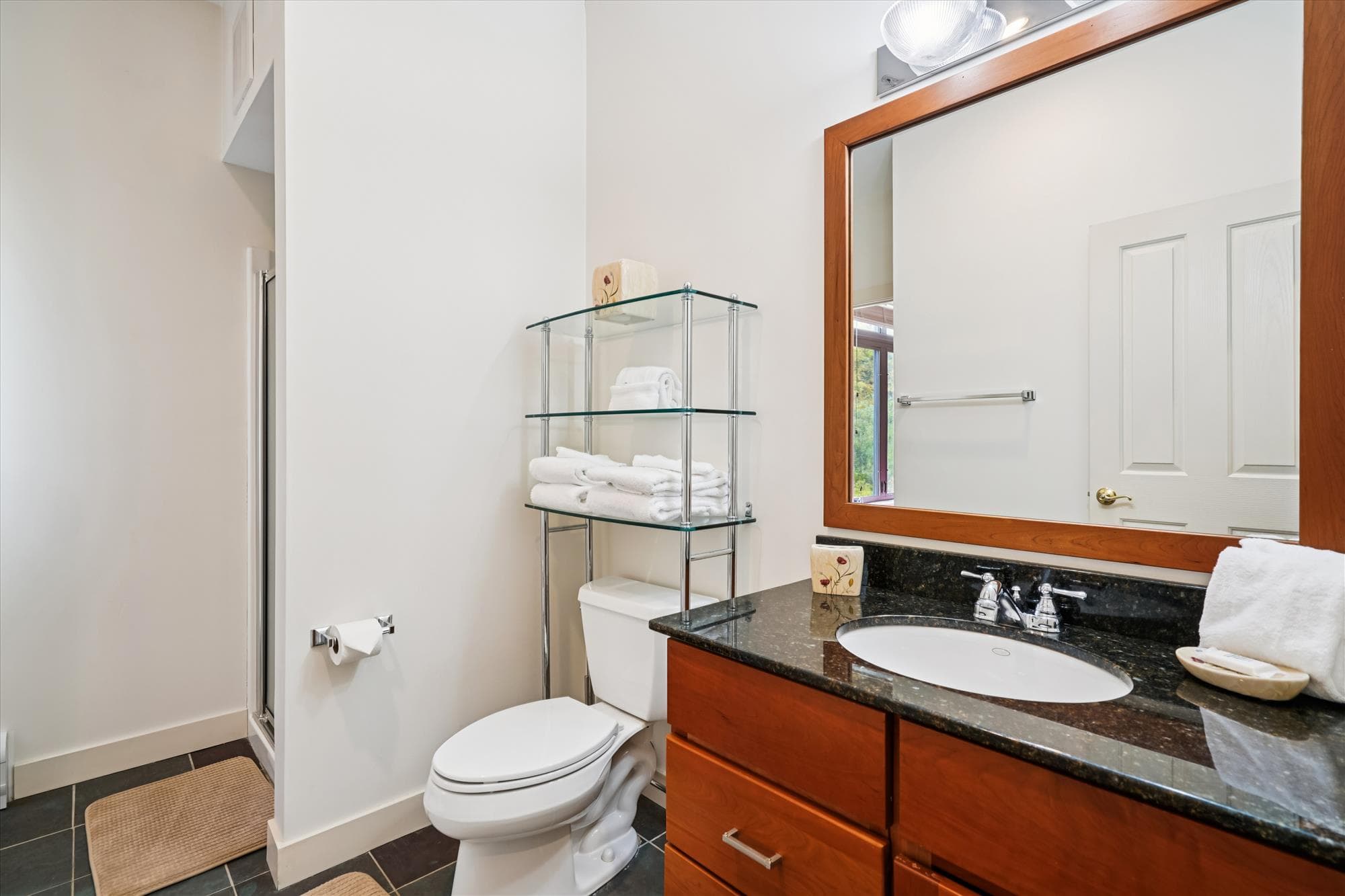 A bright full bath featuring a walk-in shower, granite vanity, and glass shelving stocked with fresh towels. Conveniently located near the queen bedroom for privacy and comfort.
