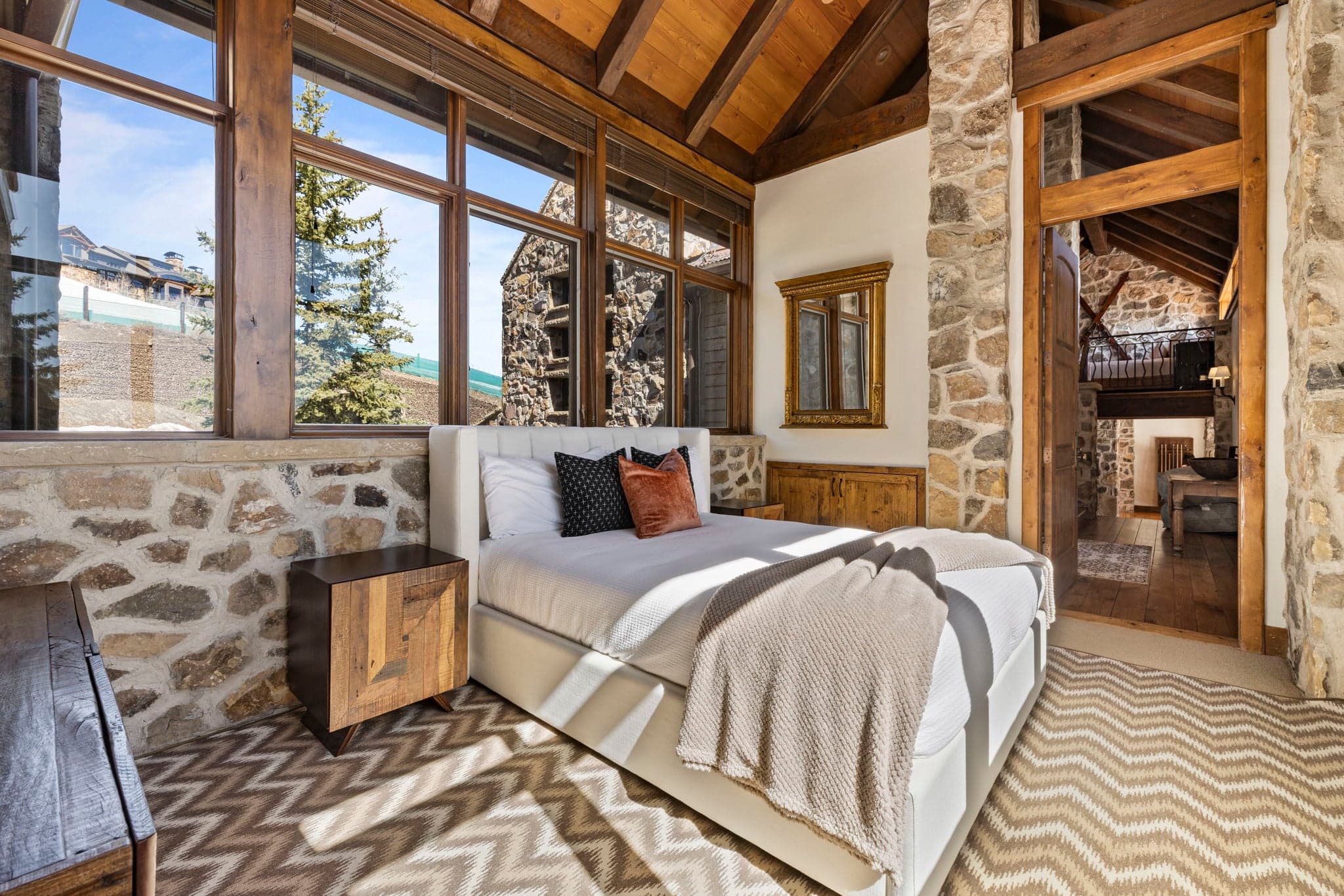 Guest suite with cozy upholstered bed, vaulted wood ceiling, and stone accent wall.