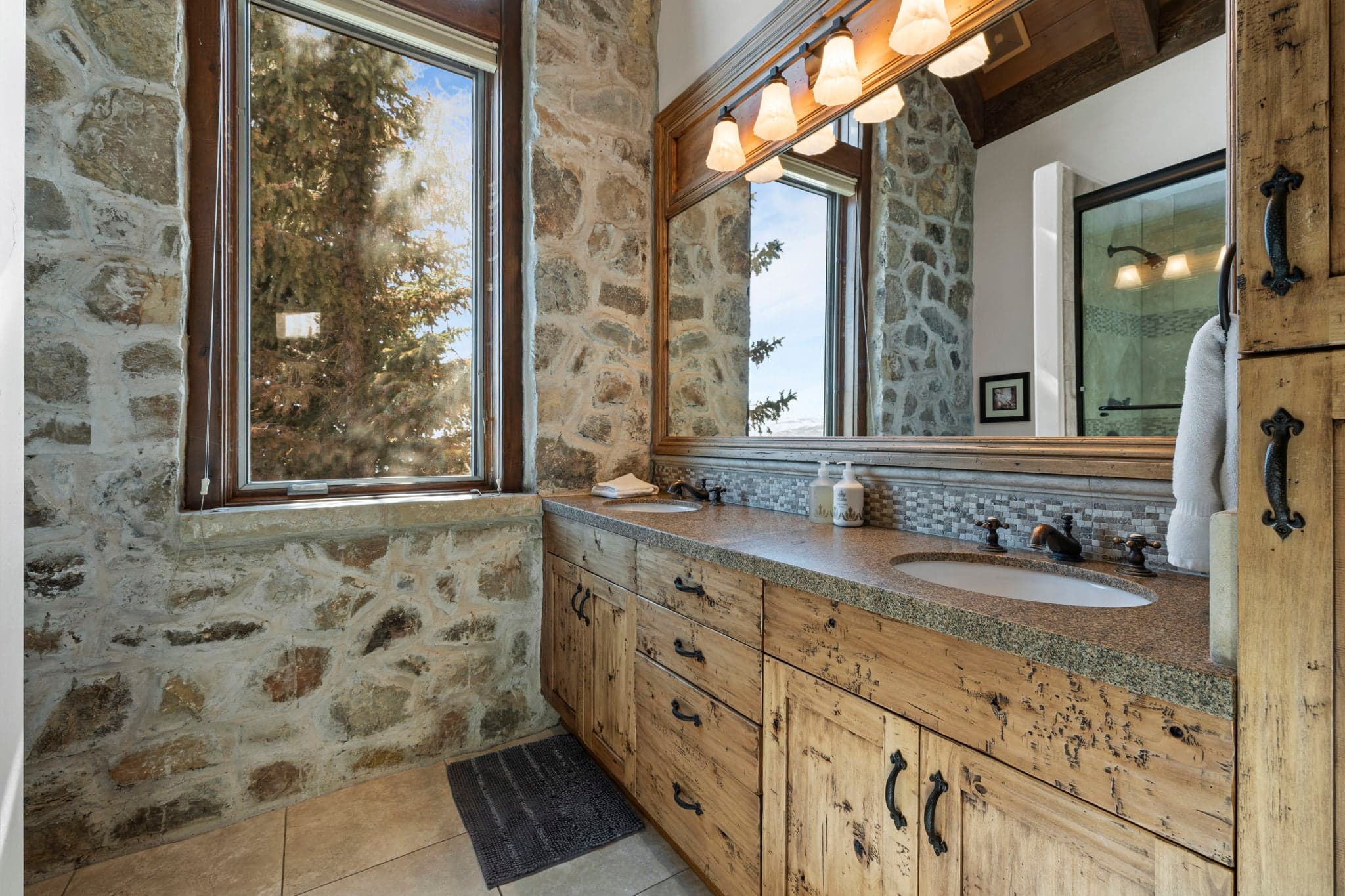 Bathroom with dual-sink wood vanity, stone walls, and large mirror.