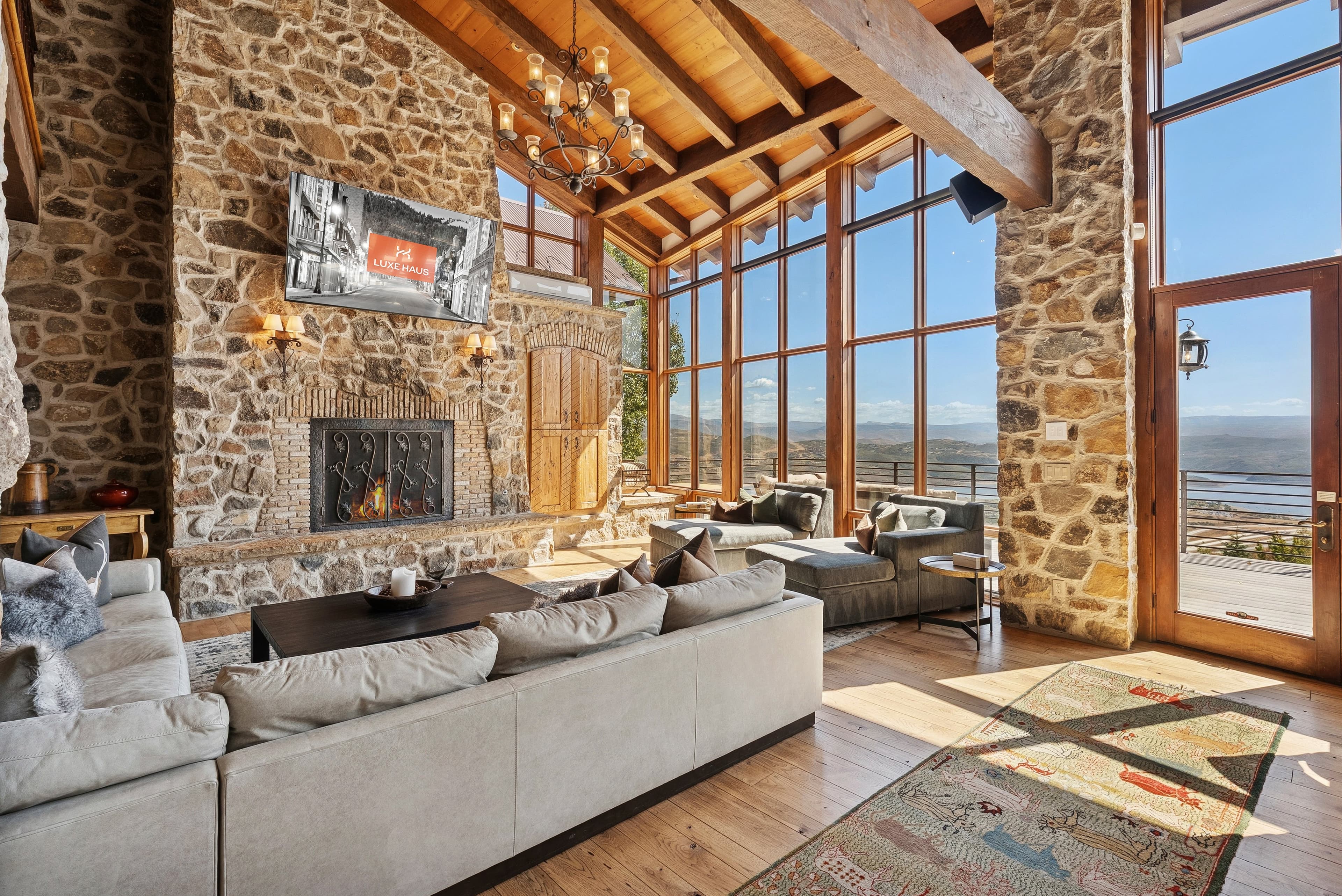 Living room with stone fireplace, vaulted wood ceiling, and floor-to-ceiling windows with mountain views.